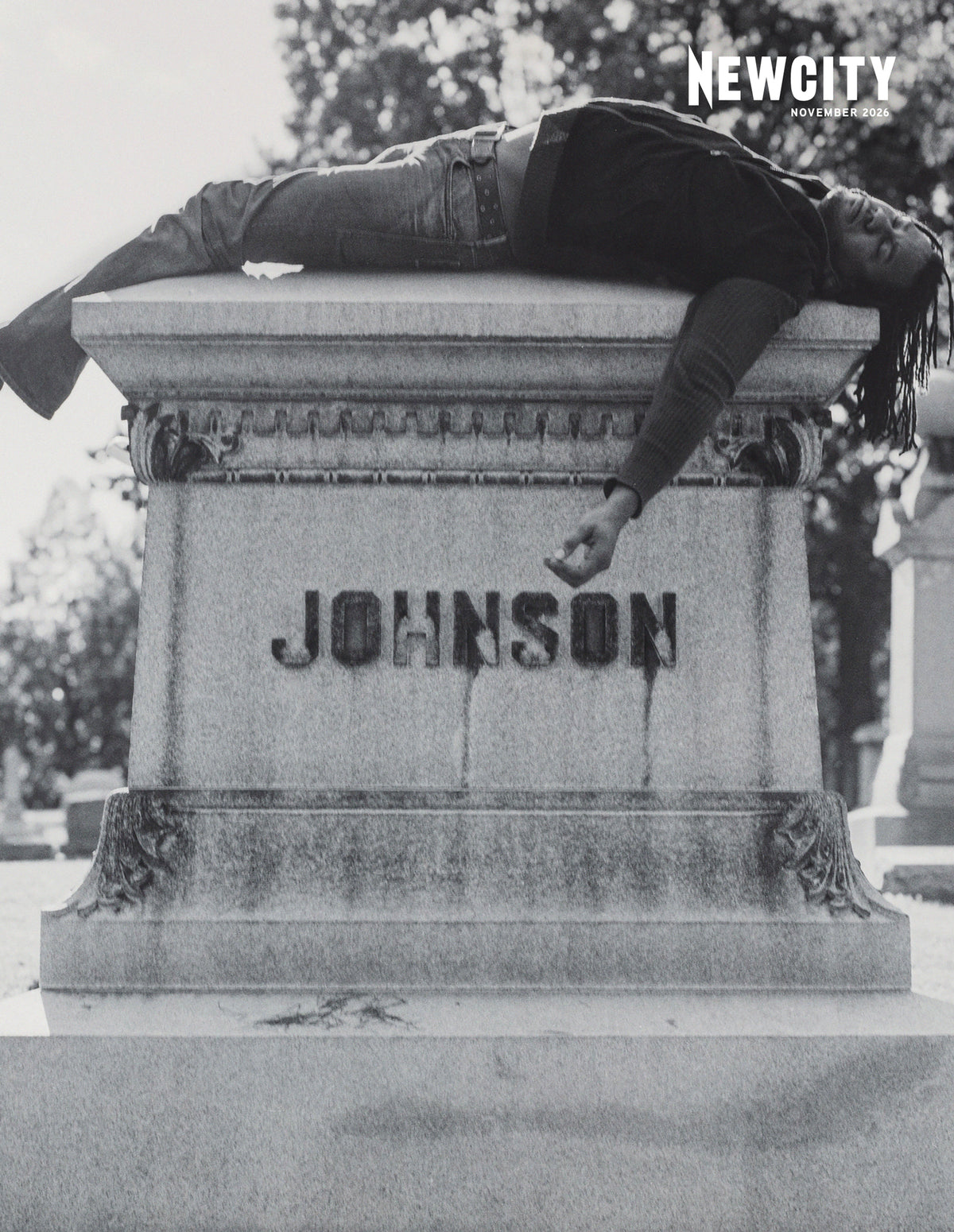A person with long hair lies on their back on top of a stone tombstone marked JOHNSON, with one arm dangling over the edge in a style reminiscent of the November 2026 Issue: Film 50 (Print Edition).