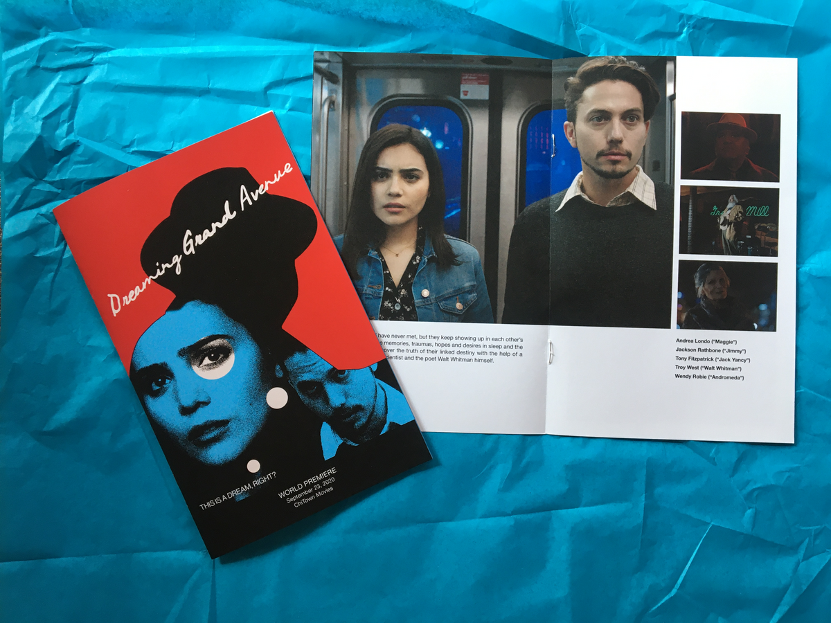 Photo of the Dreaming Grand Avenue World Premiere Program, featuring a striking red and black cover. The booklet is open to display pages with images of a woman and a man on a train, accompanied by smaller photos of additional cast members set against a blue background—an ideal keepsake for fans celebrating the world premiere.