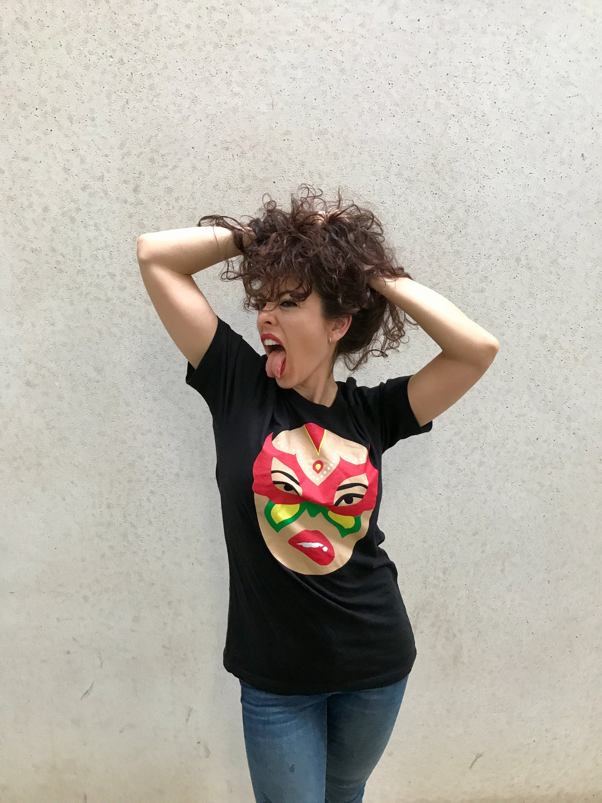 A person stands against a plain wall, wearing the Limited Edition Signature Move Wrap Party T-Shirt, which features a colorful illustrated face on the front. They playfully pull their curly hair with both hands and stick their tongue out.