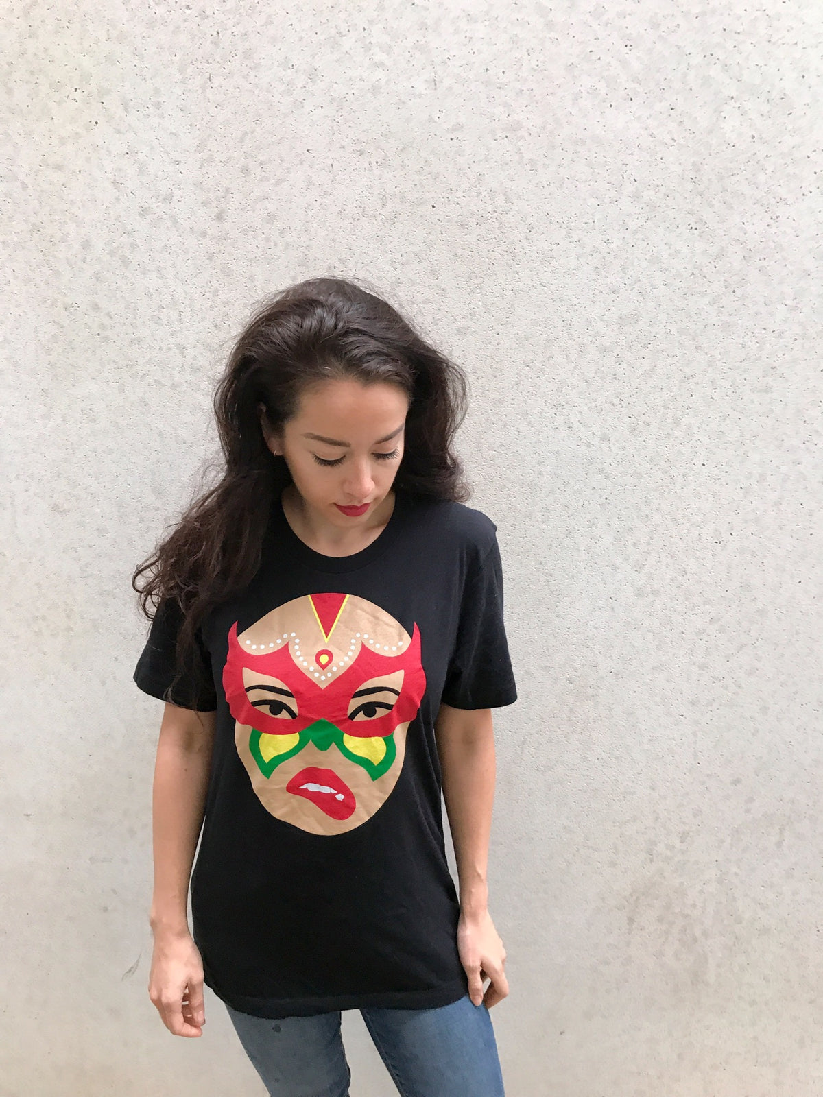 A woman with long dark hair and red lipstick looks down while wearing the Limited Edition Signature Move Wrap Party T-Shirt, which features a bold, colorful face design on the front, standing against a plain light-colored wall.