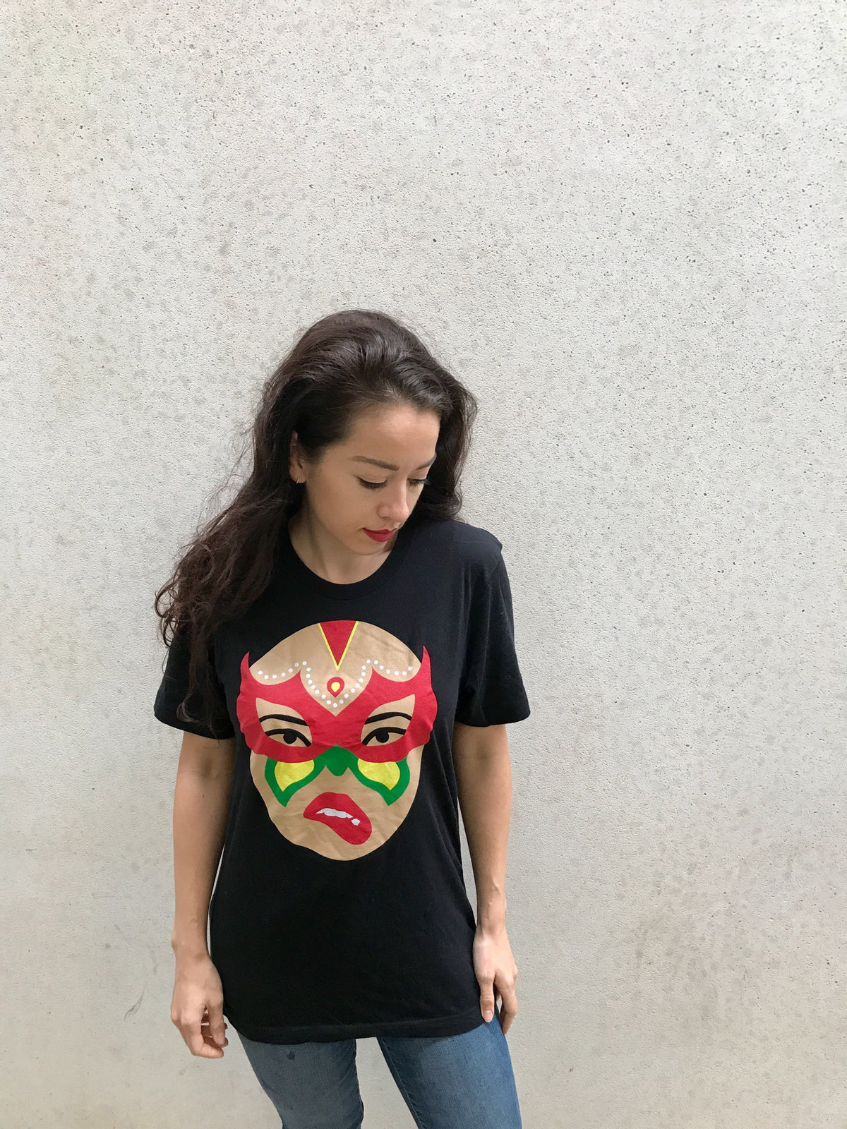 A woman with long dark hair stands against a plain light-colored wall, wearing the Limited Edition Signature Move Wrap Party T-Shirt featuring a colorful mask-like face design. She looks down and slightly to her left with one hand by her side.