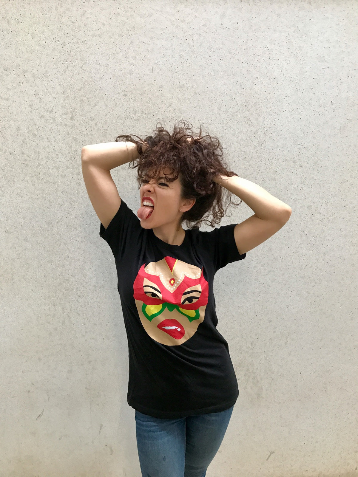 A woman stands in front of a plain wall, playfully sticking out her tongue and pulling her curly hair. She wears the Limited Edition Signature Move Wrap Party T-Shirt with a colorful cartoon face printed on it and blue jeans.