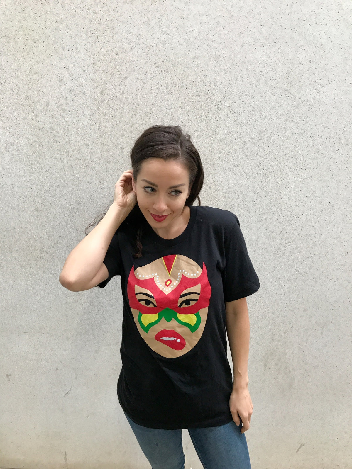 A woman with long dark hair, wearing the Limited Edition Signature Move Wrap Party T-Shirt with a vibrant mask logo graphic, stands against a plain light-colored wall, touching her hair and gazing slightly to the side.