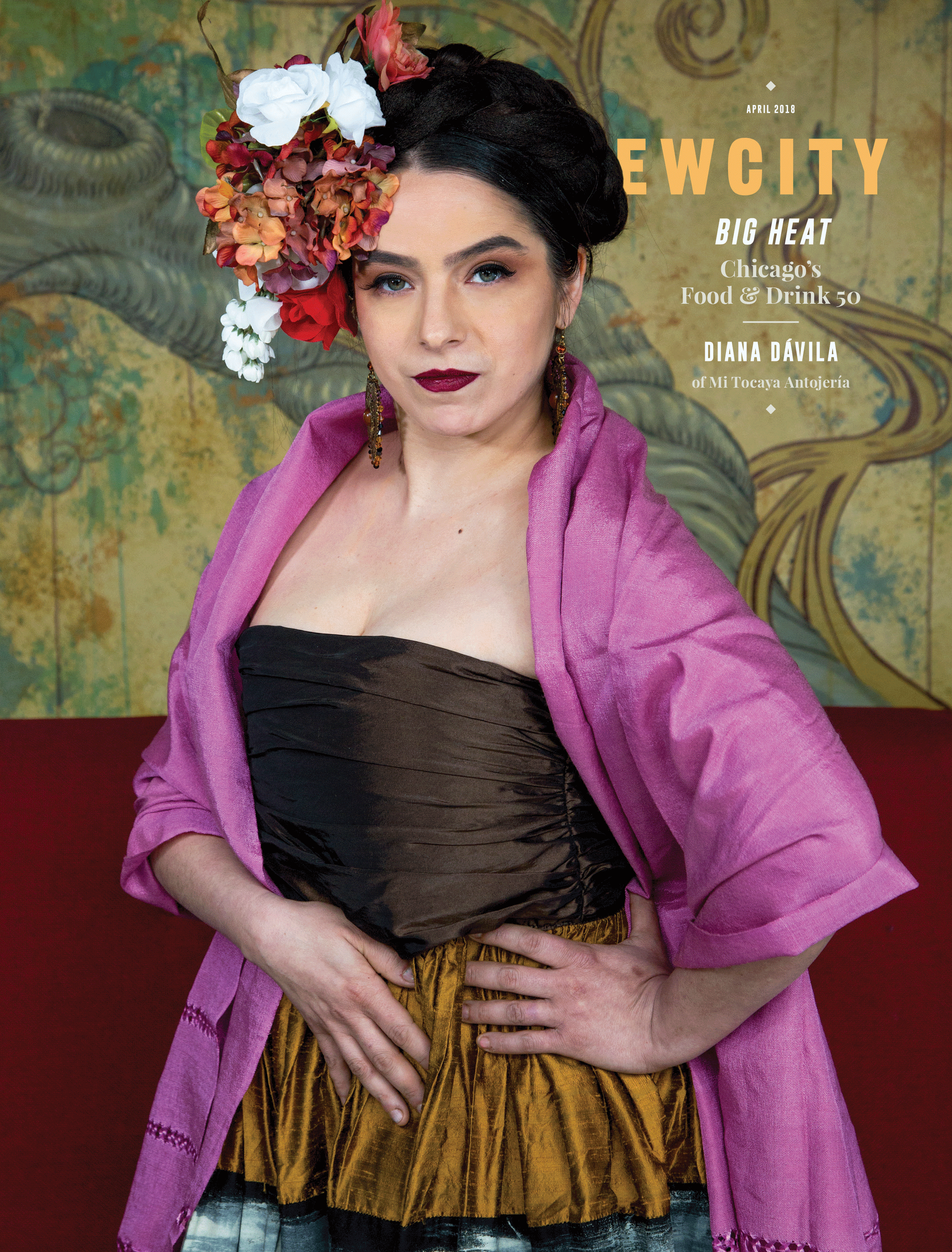 A woman with dark hair styled up and adorned with flowers poses confidently, wearing a brown dress, pink shawl, and bold makeup, standing against an ornate, artistic background that reflects the vibrant spirit showcased in the April 2018 Issue: Big Heat Food & Drink 50, celebrating Chicago’s culinary innovators like Chef Diana Dávila and other kitchen creatives.