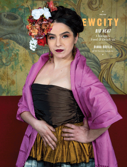A woman with dark hair styled up and adorned with flowers poses confidently, wearing a brown dress, pink shawl, and bold makeup, standing against an ornate, artistic background that reflects the vibrant spirit showcased in the April 2018 Issue: Big Heat Food & Drink 50, celebrating Chicago’s culinary innovators like Chef Diana Dávila and other kitchen creatives.