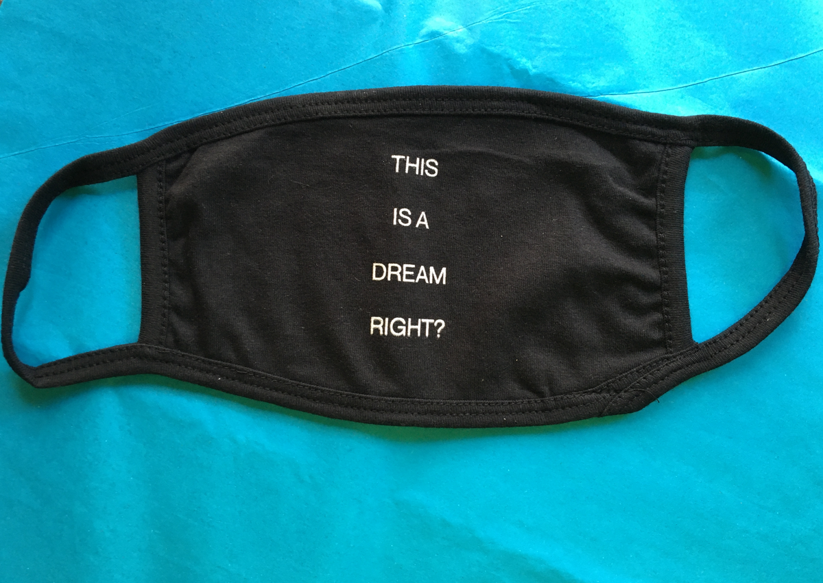 A "This is a dream, right?" face mask made of black cotton, featuring the phrase in white text and displayed flat on a blue surface—one of our commemorative designs inspired by Dreaming Grand Avenue.